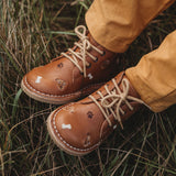Les jambes d'un enfant sur du gazon qui porte des chaussures en cuir couleur latté, avec lacets avec des détails de broderies de chiens.