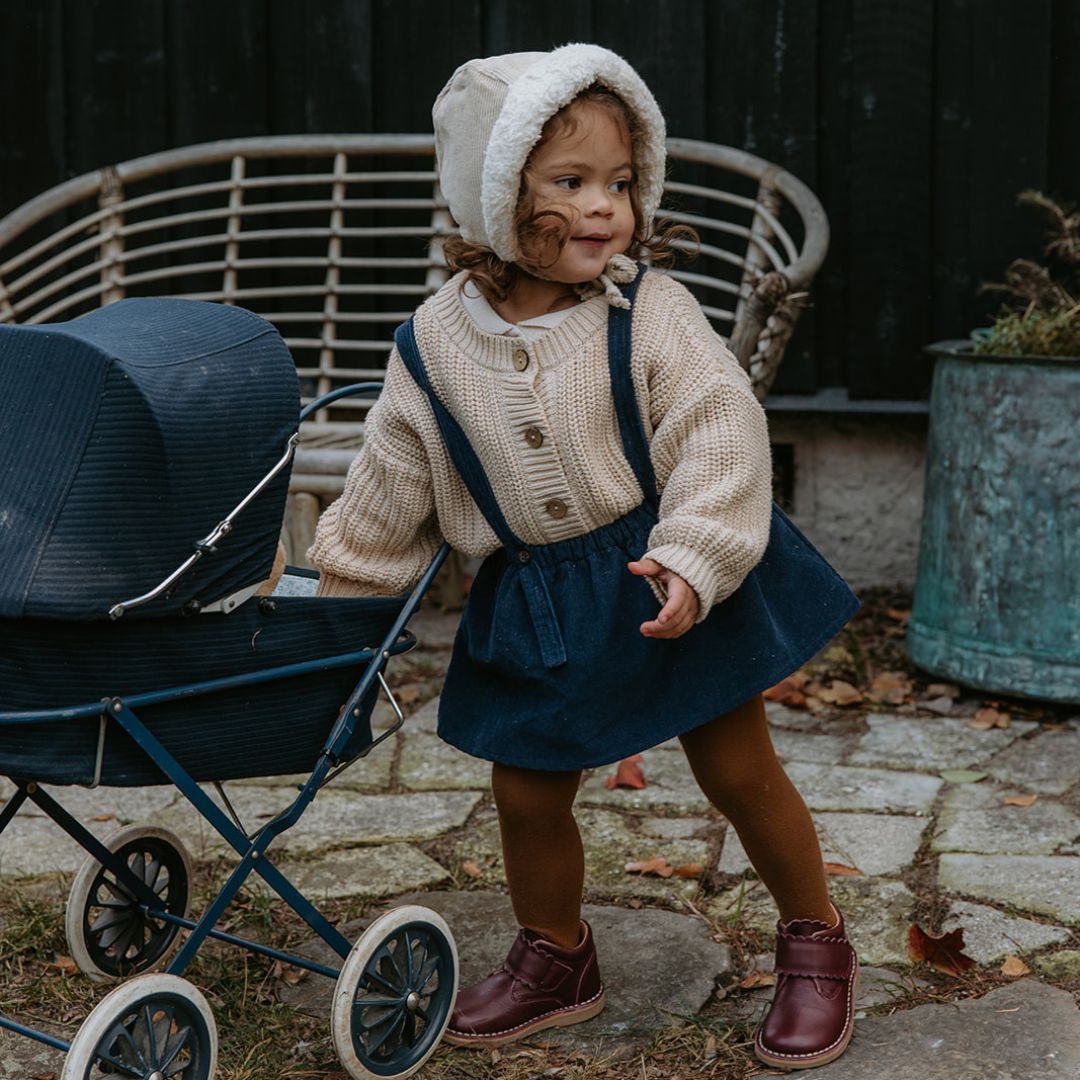 Une fillette debout jour avec une poussette et porte des chaussures Petit Nord couleur prune.
