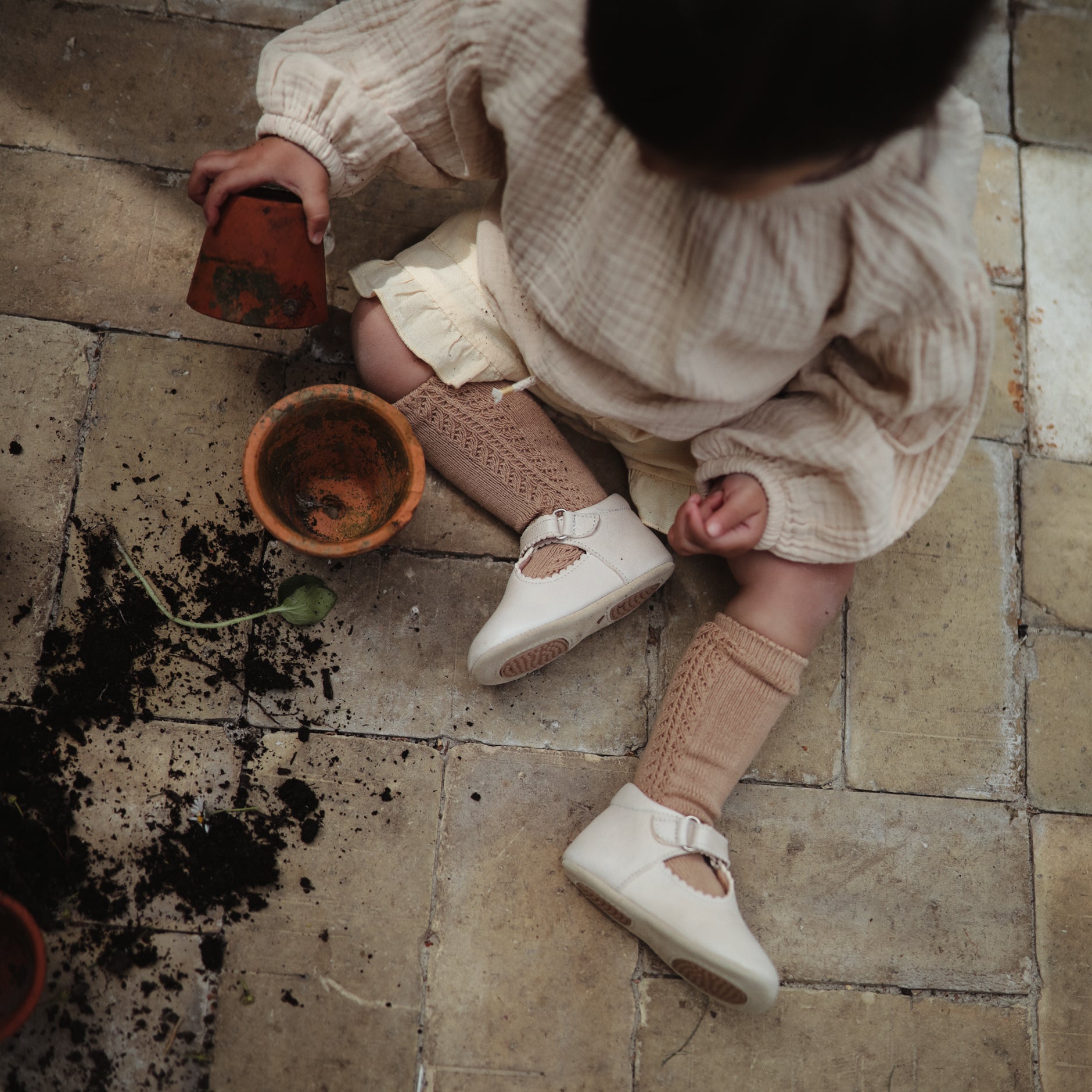Un bébé assis par terre qui joue avec la terre en portant des chaussons couleur crème. 