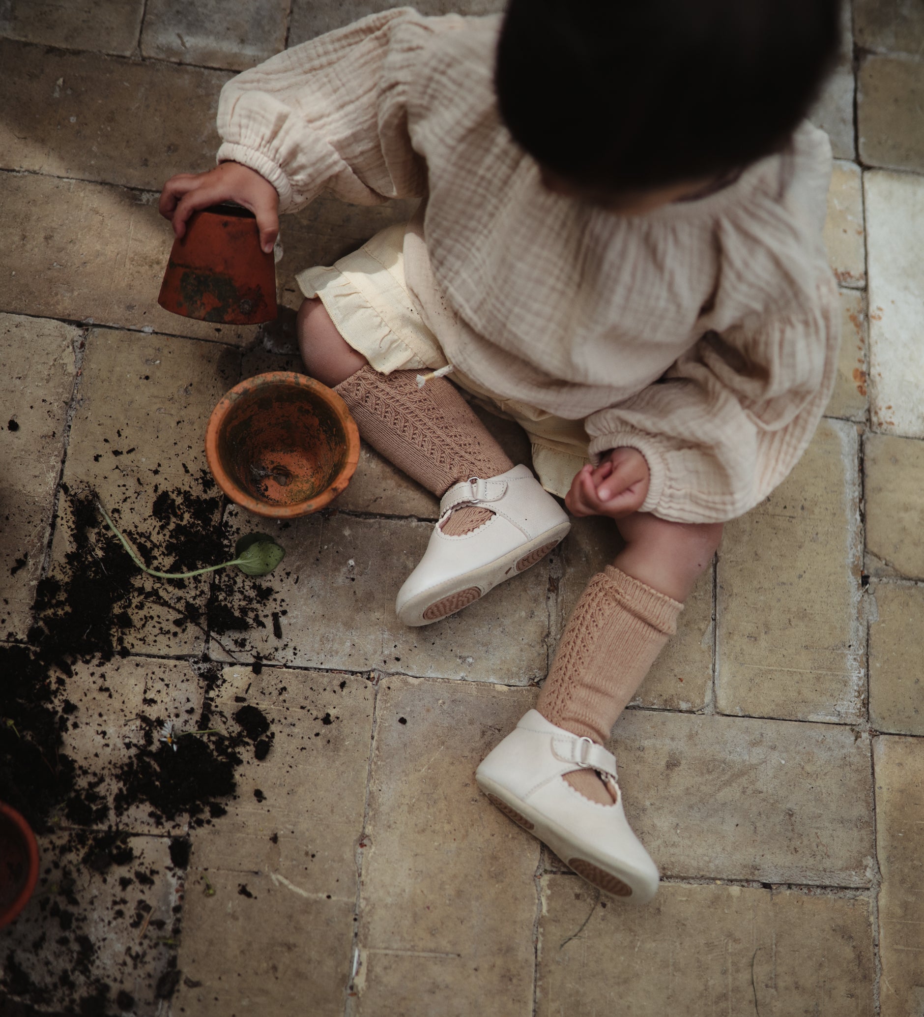 Un bébé assis par terre qui joue avec la terre en portant des chaussons couleur crème. 