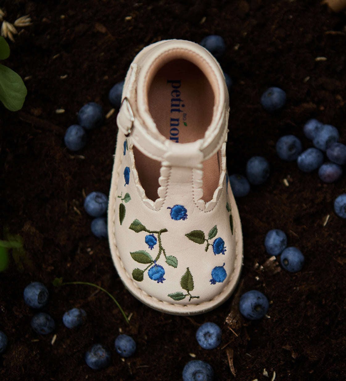 Chaussure semi-ouverte pour enfant avec détails de bleuets brodés sur de la terre à côté de véritables bleuets,