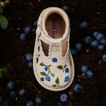 Chaussure semi-ouverte pour enfant avec détails de bleuets brodés sur de la terre à côté de véritables bleuets,