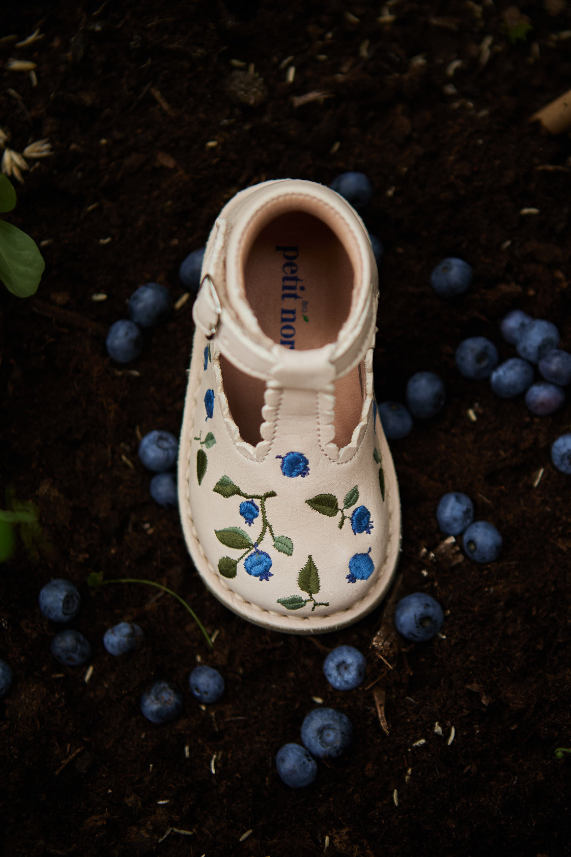 Chaussure semi-ouverte pour enfant avec détails de bleuets brodés sur de la terre à côté de véritables bleuets,