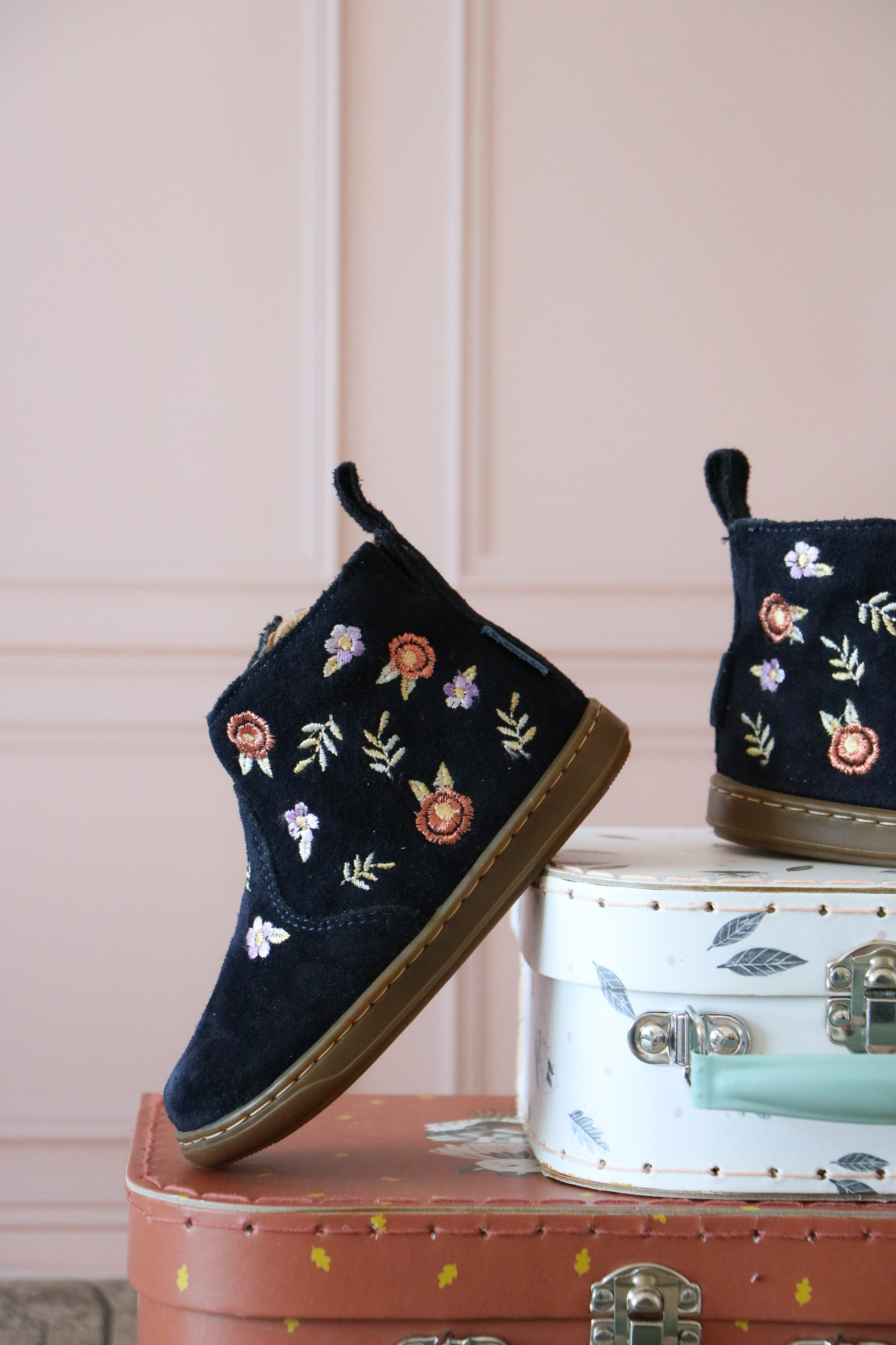 Bottine pour bébés et enfants en cuir marine avec jolis détails fleuris. Posées sur une valise d'enfant.