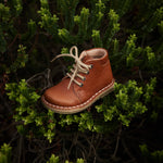 Chaussure danoise pour enfant de Petit Nord, couleur cognac, avec lacets beiges, look vintage. Déposée dans un arbuste.