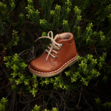 Chaussure danoise pour enfant de Petit Nord, couleur cognac, avec lacets beiges, look vintage. Déposée dans un arbuste.
