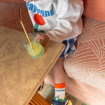Un garçon de 3 ans assis sur une banquette porte des shorts et chaussures Shoo Pom colorées