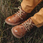 Les jambes d'un enfant sur du gazon qui porte des chaussures en cuir couleur latté, avec lacets avec des détails de broderies de chiens.