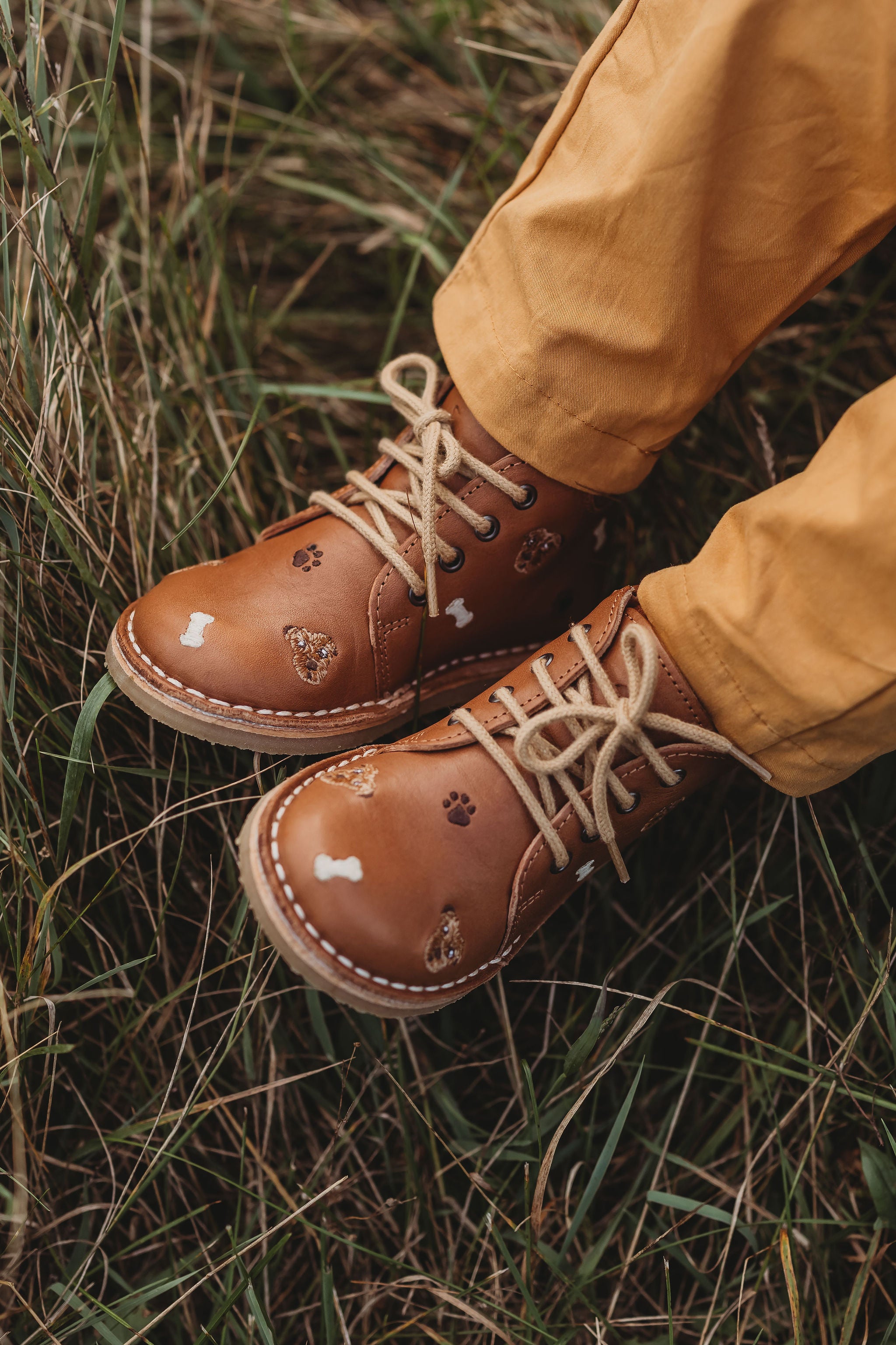 Les jambes d'un enfant sur du gazon qui porte des chaussures en cuir couleur latté, avec lacets avec des détails de broderies de chiens.
