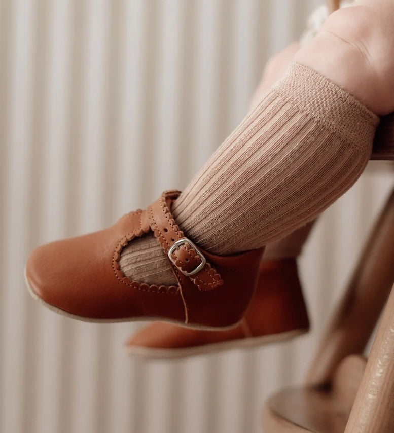 Un bébé assis porte des chaussons souples Petit Nord couleur cognac, de type semi-ouverts.